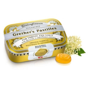 Gelbe Dose mit Grether's Pastillen für Hals und Stimme, umgeben von Holunderblüten.